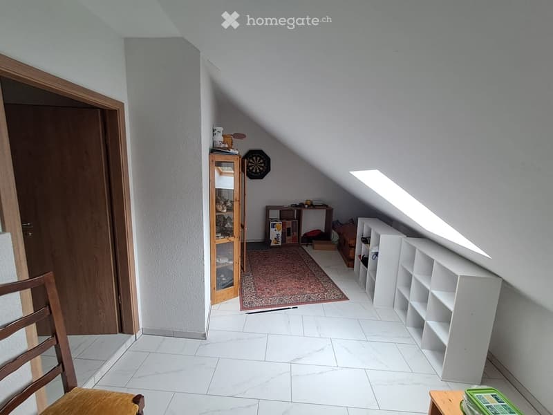 Bright attic room with white tiled flooring, slanted ceilings, and large skylight. Contains a wooden chair, glass-fronted cabinet, white shelving unit, small table, and dartboard on the wall.