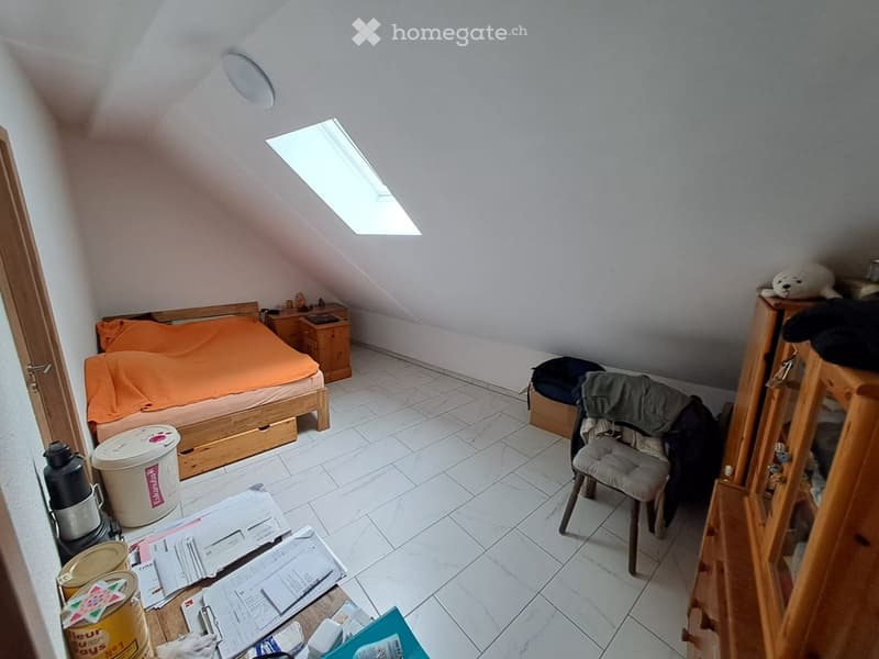 A small, slanted-ceiling bedroom with a single bed, nightstand, chair, and various items scattered on the floor.