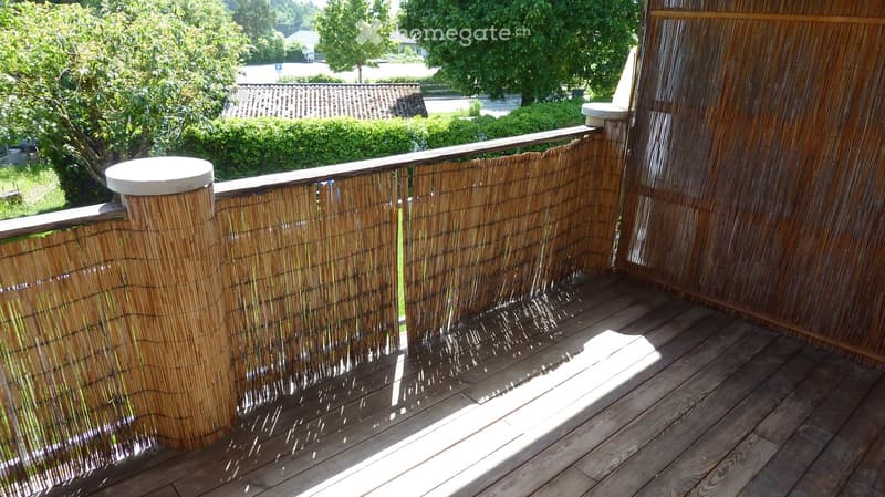 Wooden balcony with bamboo privacy screens and a round table, overlooking a green garden area.