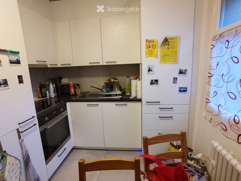 Modern kitchen with white cabinets, black countertops, and stainless steel appliances including a refrigerator, oven, and sink. A wooden chair is visible near the refrigerator.