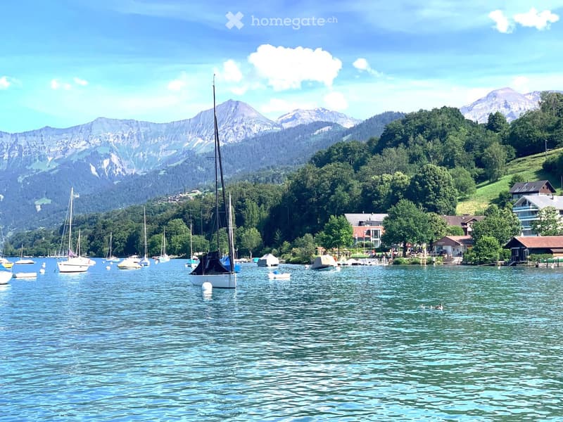 A serene lakeside scene with sailboats docked, surrounded by lush green trees and distant snow-capped mountains under a clear blue sky.