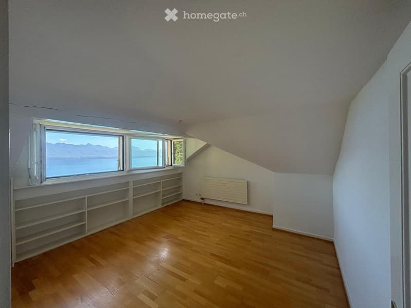 Bright, empty attic room with wooden flooring, built-in shelving, large windows offering a scenic view, and a radiator on one wall.