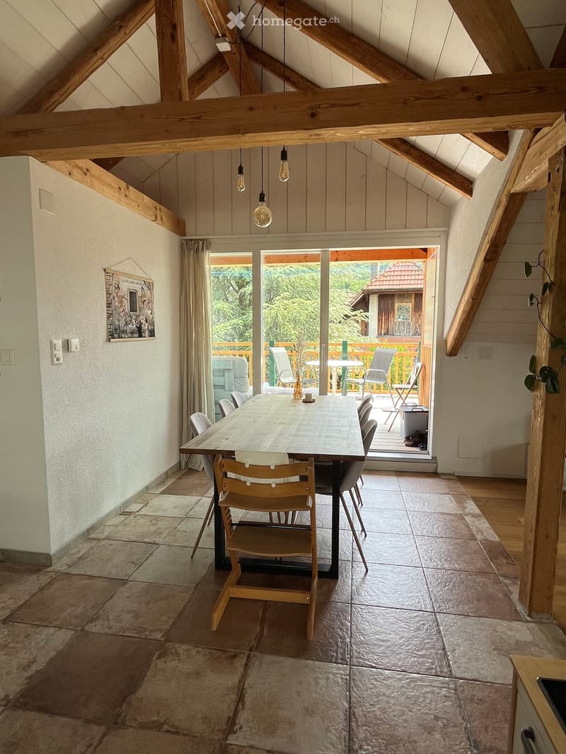 Dining area with wooden beams, large wooden dining table, chairs, and a view to a balcony with outdoor furniture.