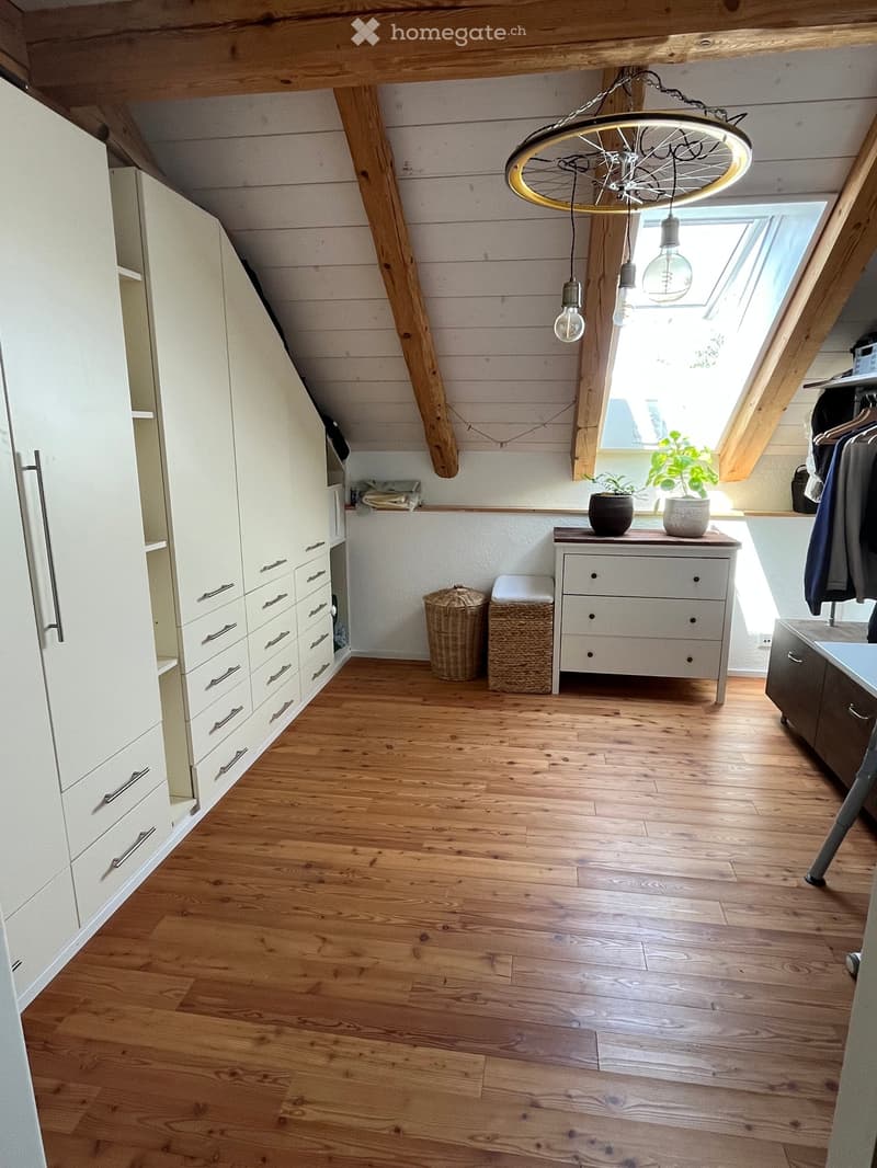 Bright attic room with wooden floors, slanted wooden ceiling beams, large skylight window, white wardrobe, small drawer unit, two stools, wicker baskets, and potted plants.