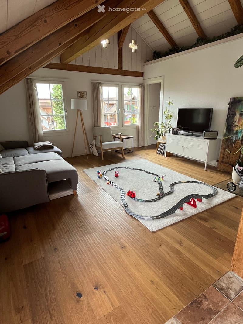 Bright living room with wooden floors, large windows, a grey sofa, armchair, coffee table, TV cabinet, floor lamp, and a toy train set on a white rug.