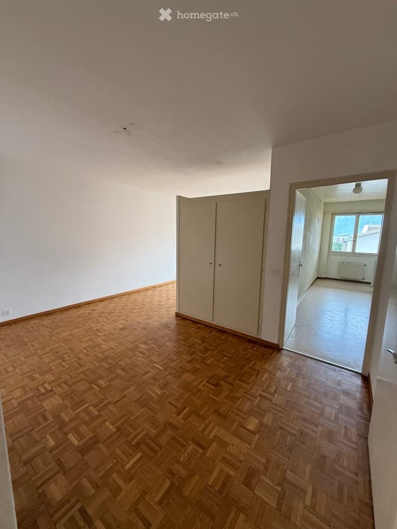 Empty room with wooden herringbone flooring, white walls, and a built-in closet. An open doorway leads to another unfurnished space with a window and radiator.