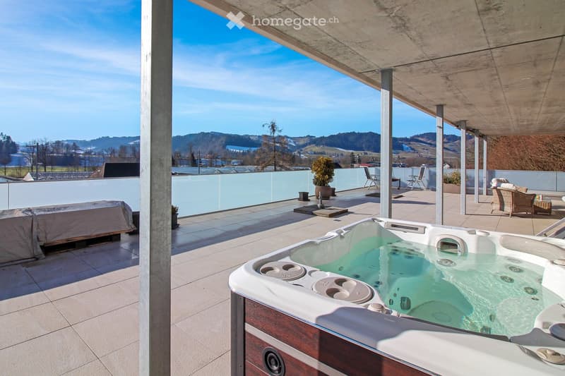 Modern rooftop terrace with a hot tub, lounge chairs, a small table, and potted plants, offering scenic views of the surrounding landscape.