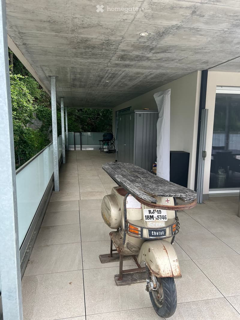 Balcony with tiled floor, scooter parked, surfboard leaning against scooter, covered area with concrete ceiling, glass railing, and outdoor furniture visible in the background.