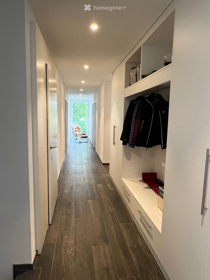 Modern hallway with white walls, tiled floor, built-in storage unit with hanging clothes, and a small seating area visible at the end.