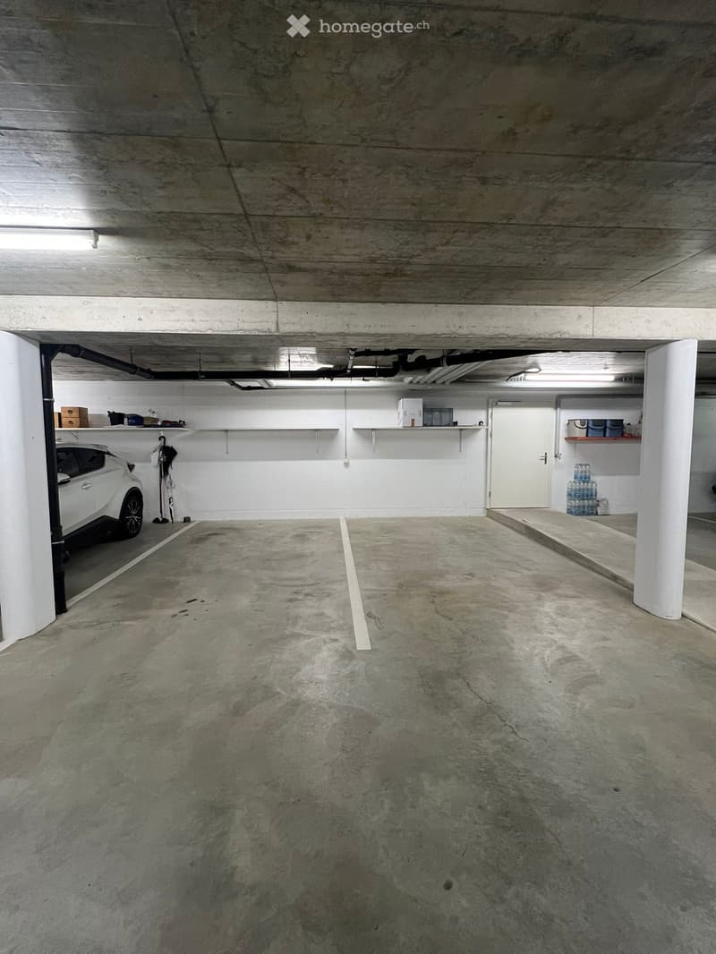 Underground parking space with a white car parked on the left, shelves with boxes and items, and a concrete floor.
