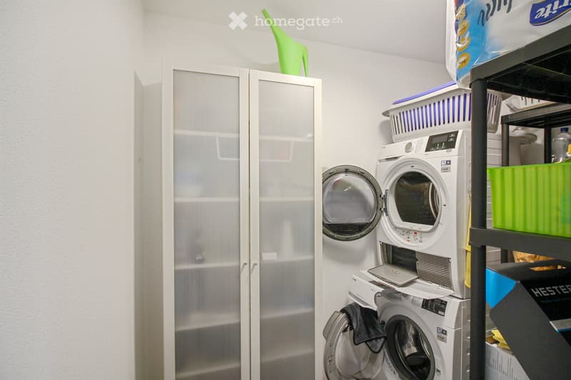 A compact bathroom with a washing machine and dryer stacked vertically, a mirrored cabinet, and a shelf with laundry baskets and cleaning supplies.