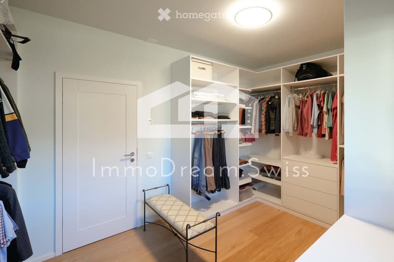 A well-organized walk-in closet with built-in white wardrobes, shelves, and a small bench. The space features wooden flooring and light-colored walls.