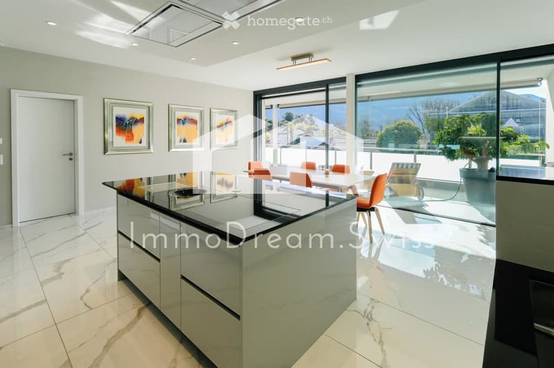 Modern kitchen with a glass island, white cabinets, and a dining area with orange chairs and a table, overlooking a scenic view through large windows.