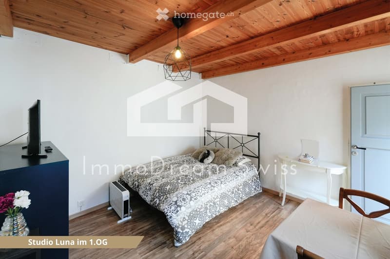 Cozy bedroom with wooden floors, exposed wooden ceiling beams, a patterned bedspread, a nightstand with a lamp, a small table, and a chair.