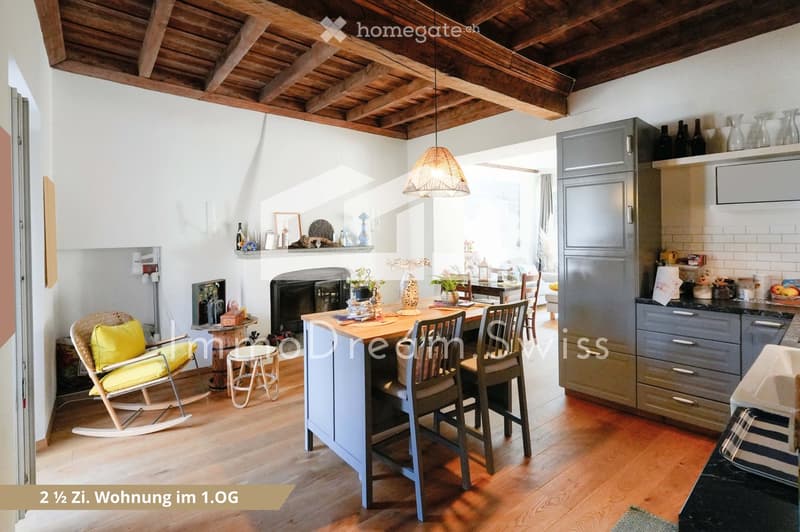 A cozy, well-furnished room with a kitchen area and a living space. Features include a wooden kitchen island with chairs, a rocking chair, a small stool, a fireplace with decorative items, and modern kitchen cabinets.