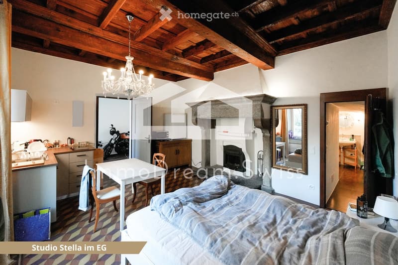 Cozy studio with exposed wooden beams, a bed, a small table and chairs, a fireplace, a kitchen area with cabinets and a counter, and a mirror. A motorcycle is visible in the adjacent room.