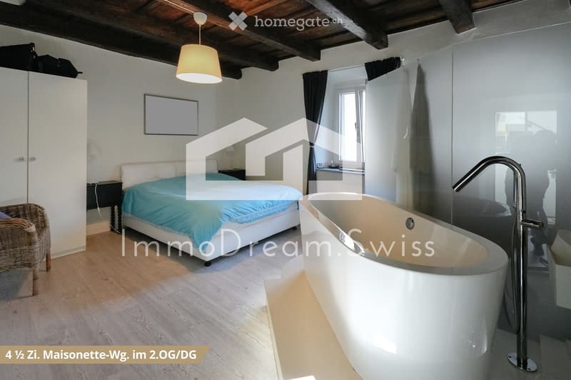 Cozy bedroom with a white bed covered in blue bedding, a wicker chair, a modern bathtub, a large mirror, and a wardrobe. The room features wooden flooring and exposed wooden beams on the ceiling.