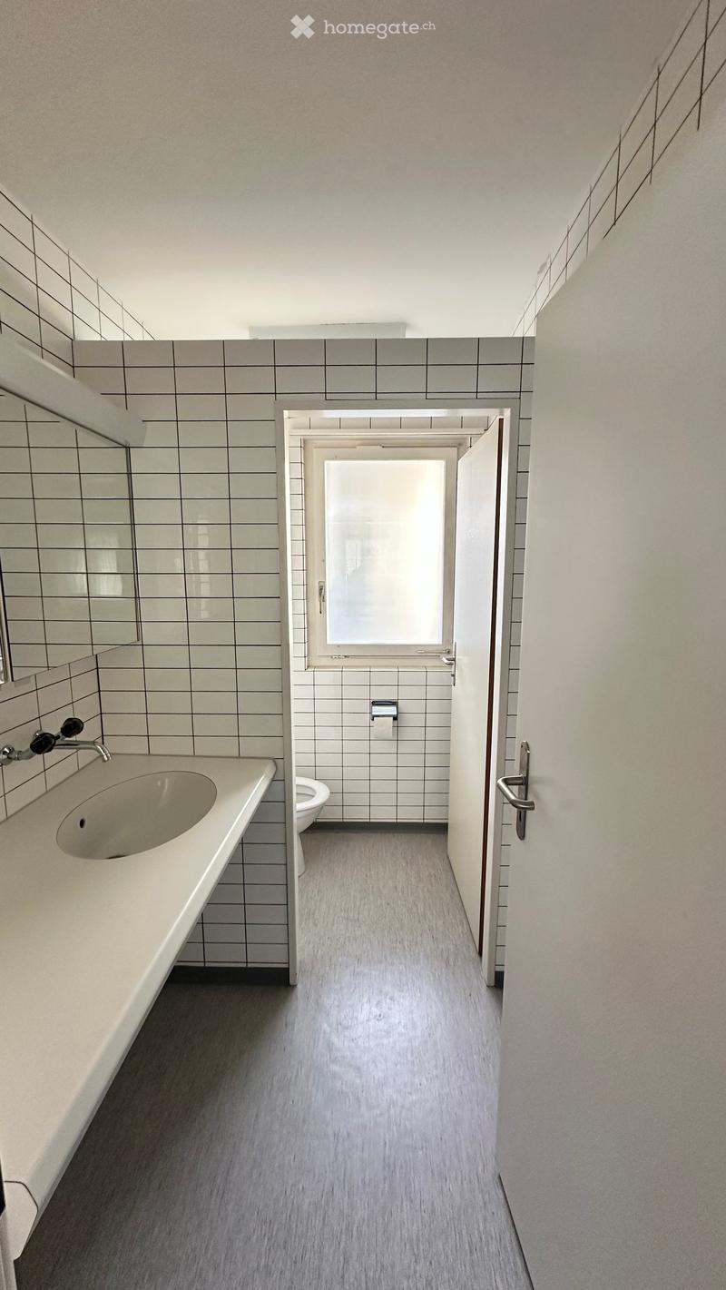 Modern bathroom with a sink, faucet, mirror, toilet, and paper towel holder. The space features grey tiled walls and a grey floor.