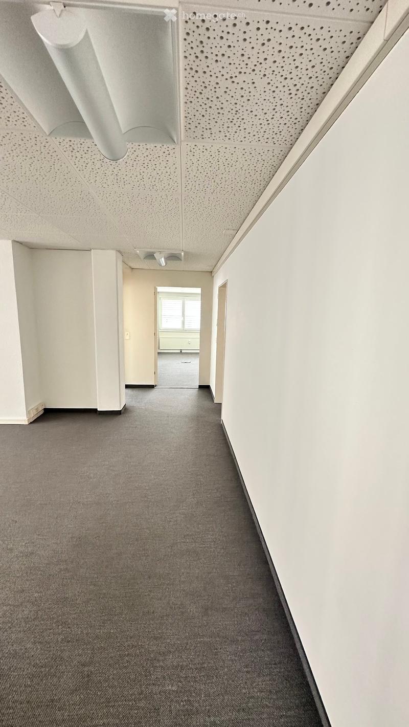 Empty hallway with grey carpet, white walls, and a perforated white ceiling. A doorway leads to another room with a window and a grey floor.