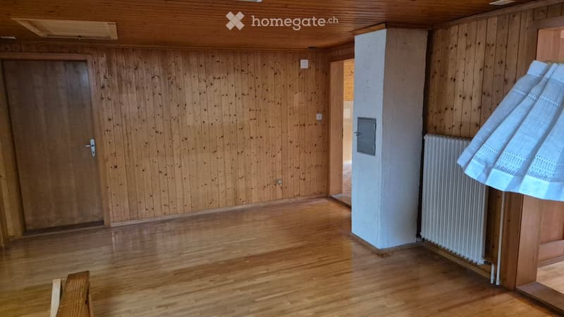 Empty room with wooden walls and floor, featuring a closed wooden door, radiator, and a partially visible cloth draped over it.