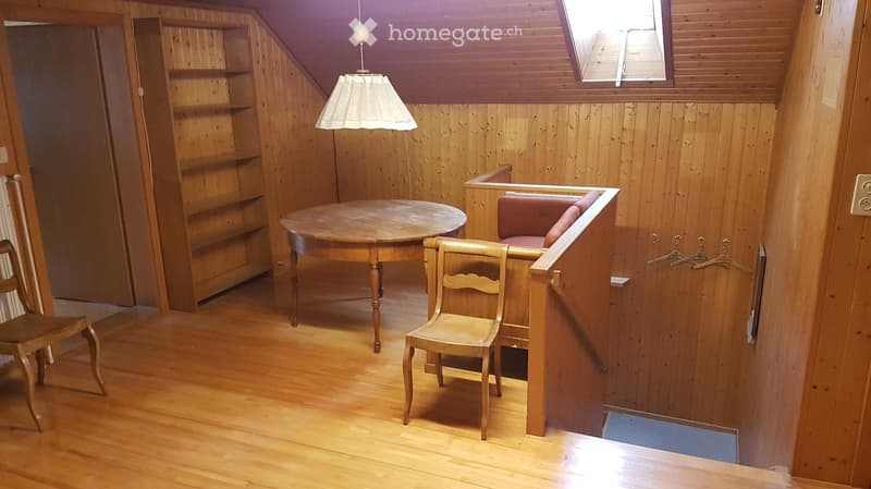 A cozy, wooden-paneled room with a round wooden table, two wooden chairs, a small sofa, a wooden shelf, and a hanging lamp.