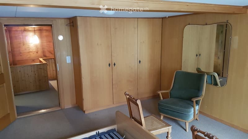 A small, furnished room with wooden walls and a grey carpet. It features a green upholstered armchair, a small wooden table, a large mirror, and a built-in wardrobe with double doors.