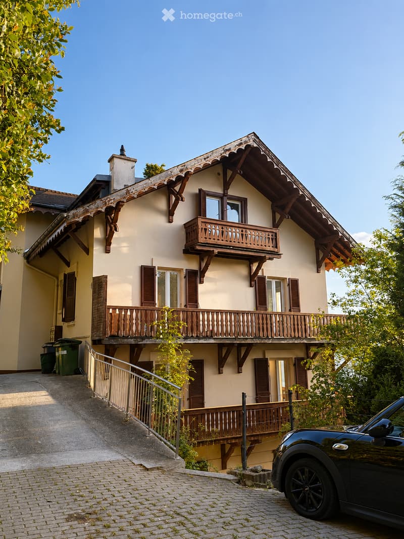 Two-story house with beige walls, wooden balconies, and multiple windows with shutters. A black car is parked on the driveway.