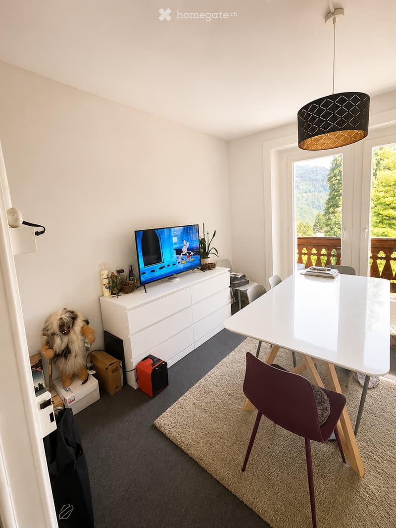 A modern, well-lit room with a white cabinet, television, dining table, chairs, and a plush toy on a stool. The room features a large window with a scenic view and a hanging lamp.
