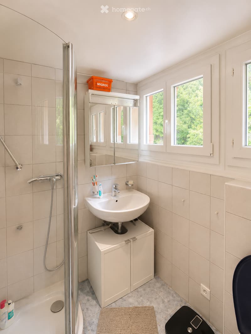 Modern bathroom with a white sink, mirror, cabinet, shower, bath, and toilet. Features a tiled wall, a small window, and a bathroom scale on the floor.