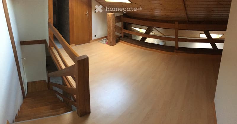 Wooden staircase with a wooden railing leads to an open loft area with exposed wooden beams and a skylight. The space is clean and well-lit, with a neutral color palette.
