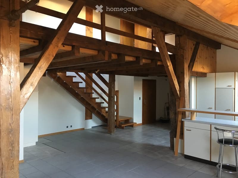 Open-plan space with exposed wooden beams, a staircase leading to an upper level, a small bench, and a kitchen area with a counter and stools.