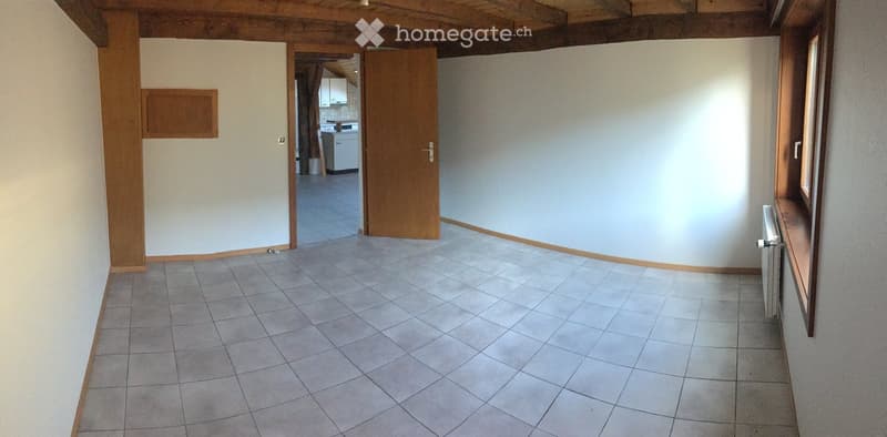 Empty room with tiled floor, white walls, wooden door frame, and a view into an adjacent kitchen area.