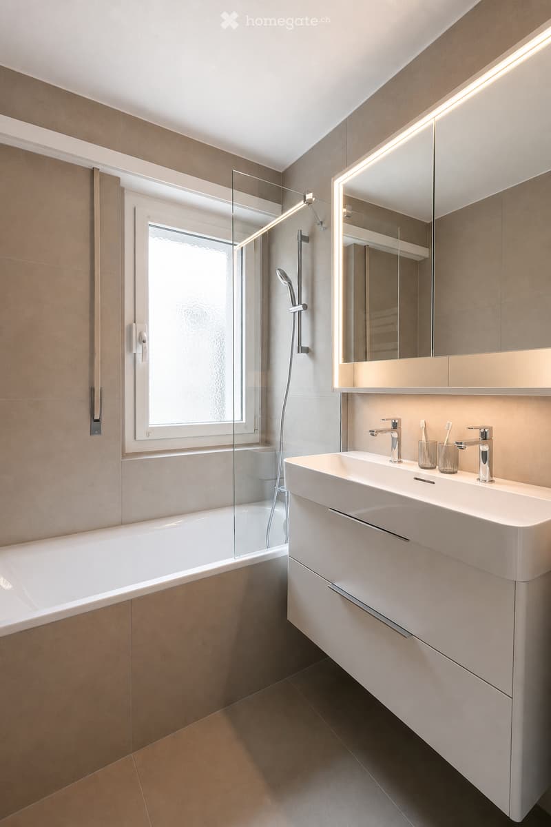 Modern bathroom with a bathtub, sink, and mirror. Features include a glass shower enclosure, built-in storage, and a sleek design with warm-toned tiles.