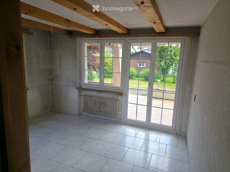 Empty room with exposed wooden beams, tiled floor, large sliding glass doors leading to a patio, radiator, and a small shelf.