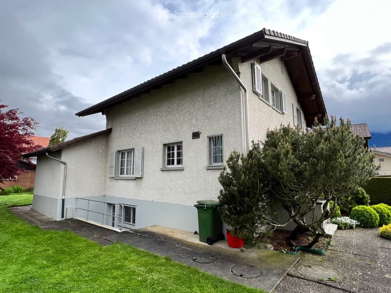 Two-story house with white textured exterior, multiple windows with shutters, and a small garden area with a tree and shrubs.