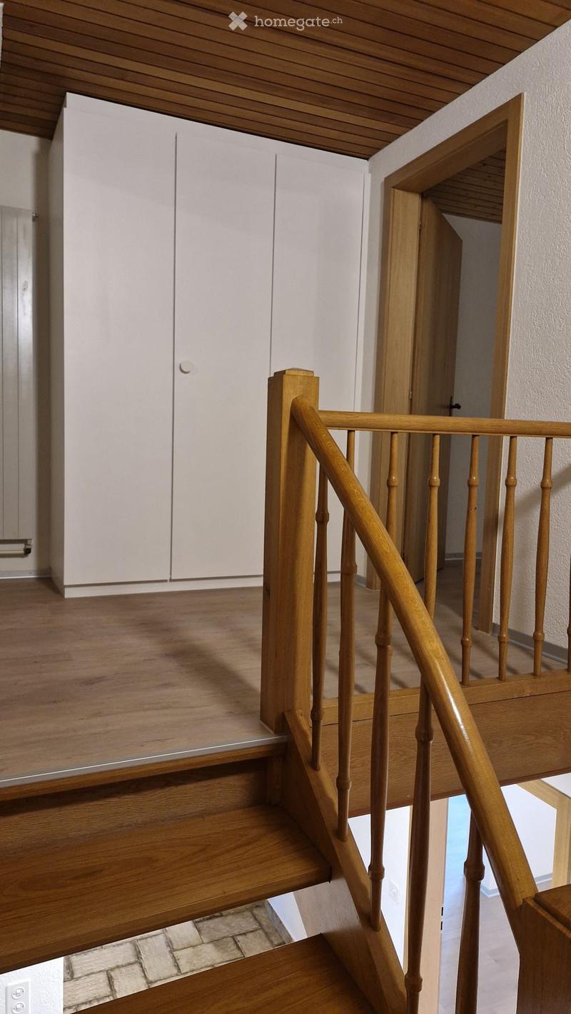 Wooden staircase with a handrail and open wooden balustrade, leading to a hallway with white walls and a wooden ceiling.