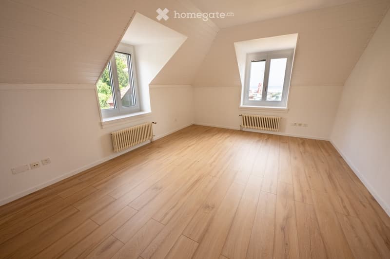 Empty attic room with wooden flooring, white walls, and two windows offering natural light. Radiators are present along the walls.