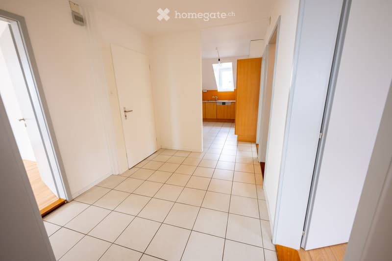 Empty hallway with tiled floor, white walls, and doors leading to other rooms, including a kitchen area with cabinets and a sink.
