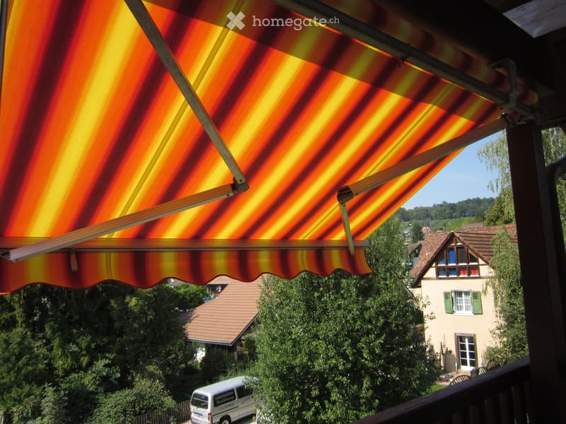 Outdoor scene with an orange and yellow awning on a balcony overlooking a house, trees, and a van parked nearby.
