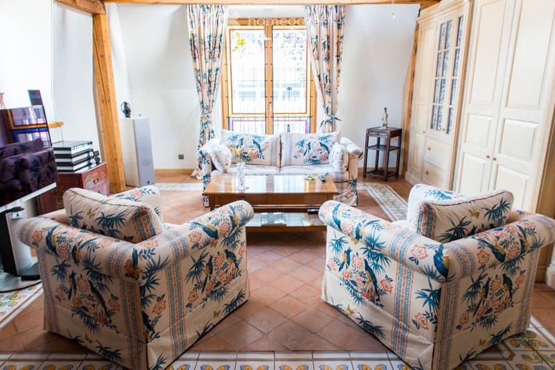 A cozy living room with a patterned sofa, two armchairs, a coffee table, side table, bookshelf, and large windows with curtains.