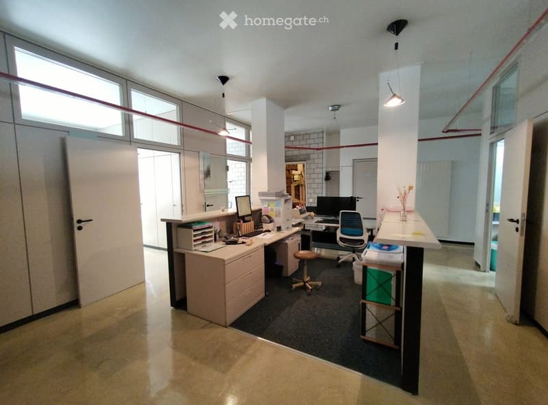 Modern office space with desks, chairs, computers, and a printer. Large windows provide natural light, and the room features a mix of carpeted and tiled flooring.