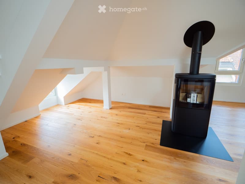 Bright, empty attic room with wooden flooring, white walls, and a modern black wood-burning stove with a chimney. Large windows allow natural light to fill the space.
