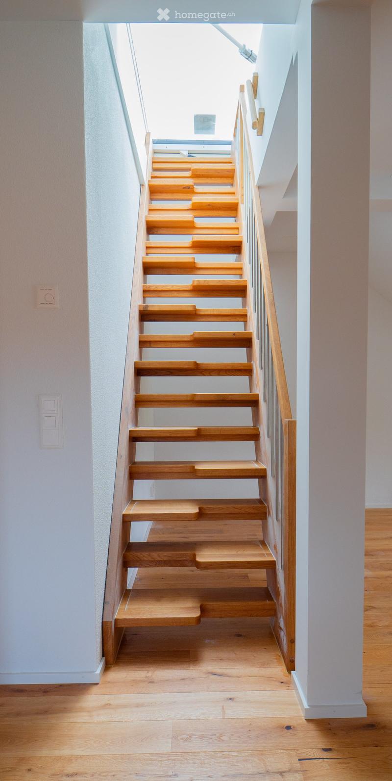 Wooden staircase with light-colored steps and a wooden handrail, set against white walls and a wooden floor.