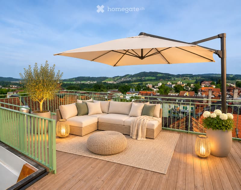 A modern rooftop terrace with a wooden deck, green railing, and a panoramic city view. Features a beige sectional sofa, ottoman, woven rug, umbrella, and decorative lamps.