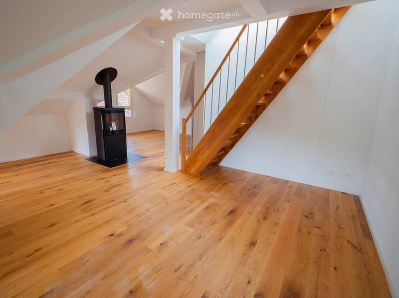 Empty attic space with wooden flooring, white walls, and a staircase leading upwards. A black wood-burning stove is positioned against the wall.