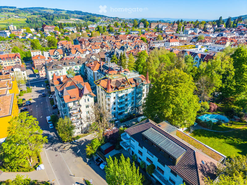 Aerial view of a residential area with multiple buildings, red-tiled roofs, green spaces, and a swimming pool in the foreground.