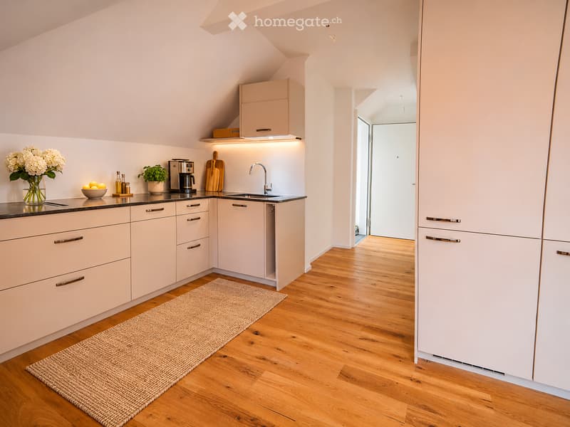 Modern kitchen with light-colored cabinets, wooden flooring, and a small rug. Features a sink, coffee maker, cutting board, vase with flowers, and a potted plant on the counter.