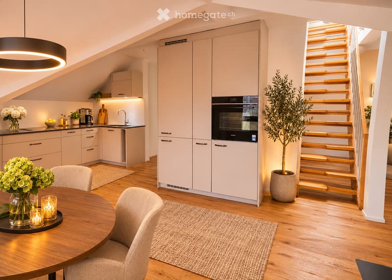 Modern kitchen and dining area with light-colored cabinets, a round wooden dining table, four chairs, a pendant light, and a staircase leading upwards.