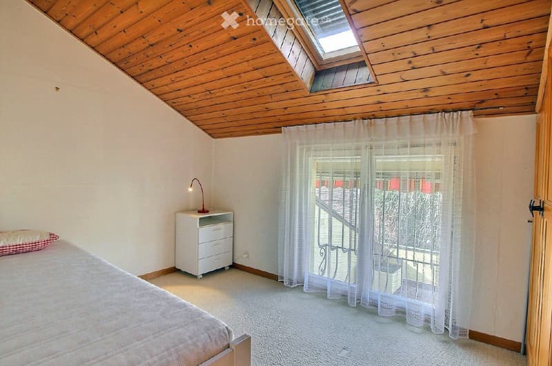 Cozy bedroom with a wooden slanted ceiling, skylight window, bed with a pillow, nightstand with a lamp, and sheer curtains covering a window with a view of a staircase.