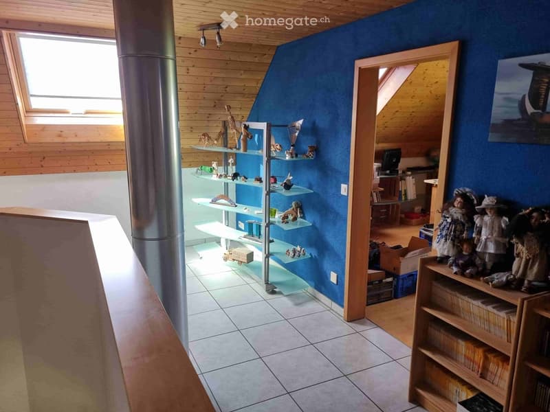A well-lit room with wooden walls, glass shelves filled with decorative items, dolls, books, and a computer screen. The room features a slanted ceiling and skylights.